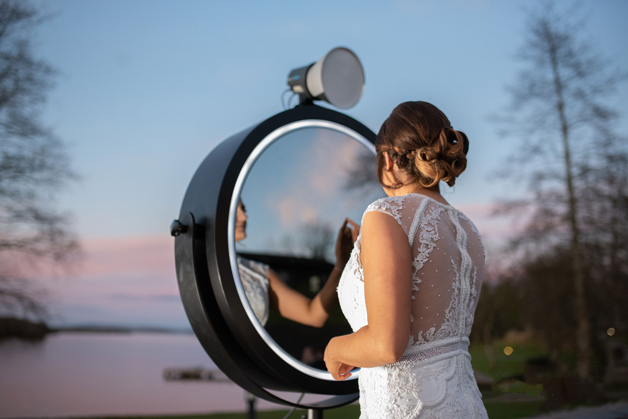 Brud i vit klänning som poserar framför en Magic Mirror-fotobås vid en sjö, vilket skapar